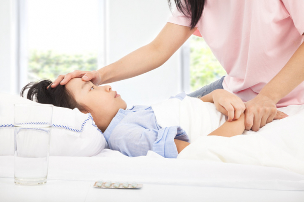 A child with dark hair lying in bed looking sick, mother in pink shirt has one hand on his forehead, the other on his hand